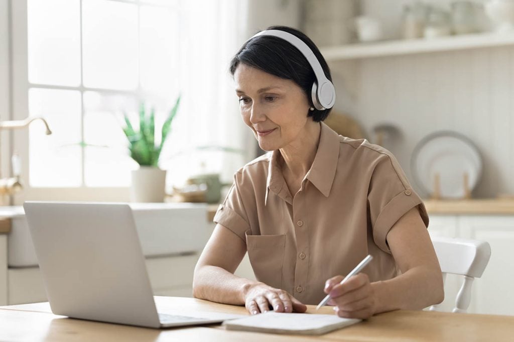 Middle-aged woman in headphones sit at table in kitchen look at laptop screen