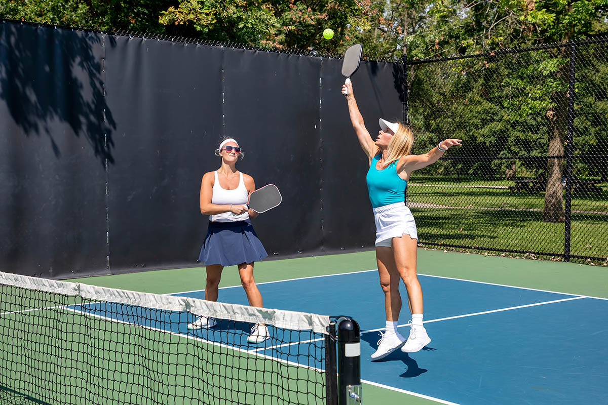 A female pickleball player returns a high pop up as her partner looks on.