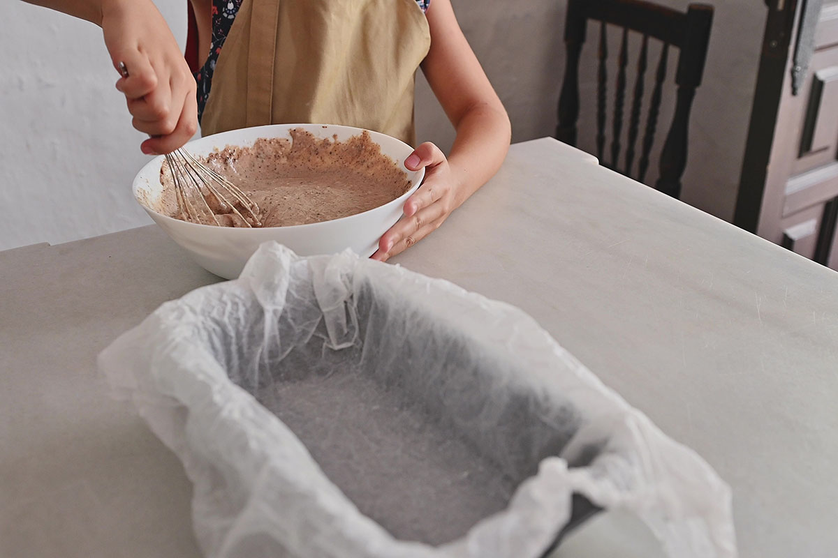 Close-up of a person mixing brownie batter in a white bowl on a kitchen counter, with a lined baking pan ready nearby. Home baking and preparation concept.