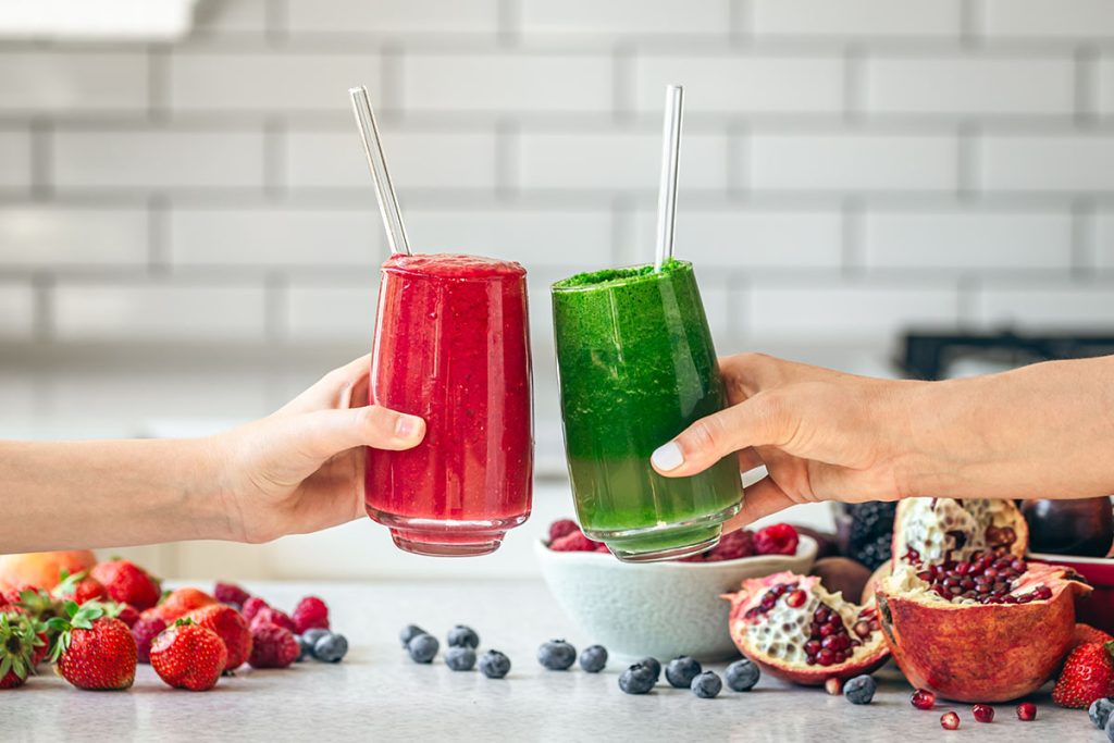 Glasses with green and red smoothies in female hands on the kitchen table.