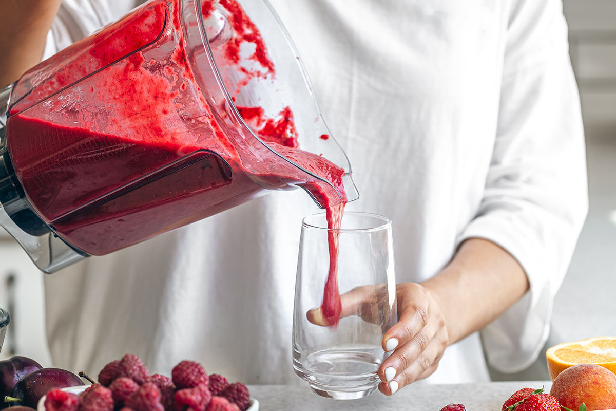 A woman pours a berry smoothie from a blender into a glass.