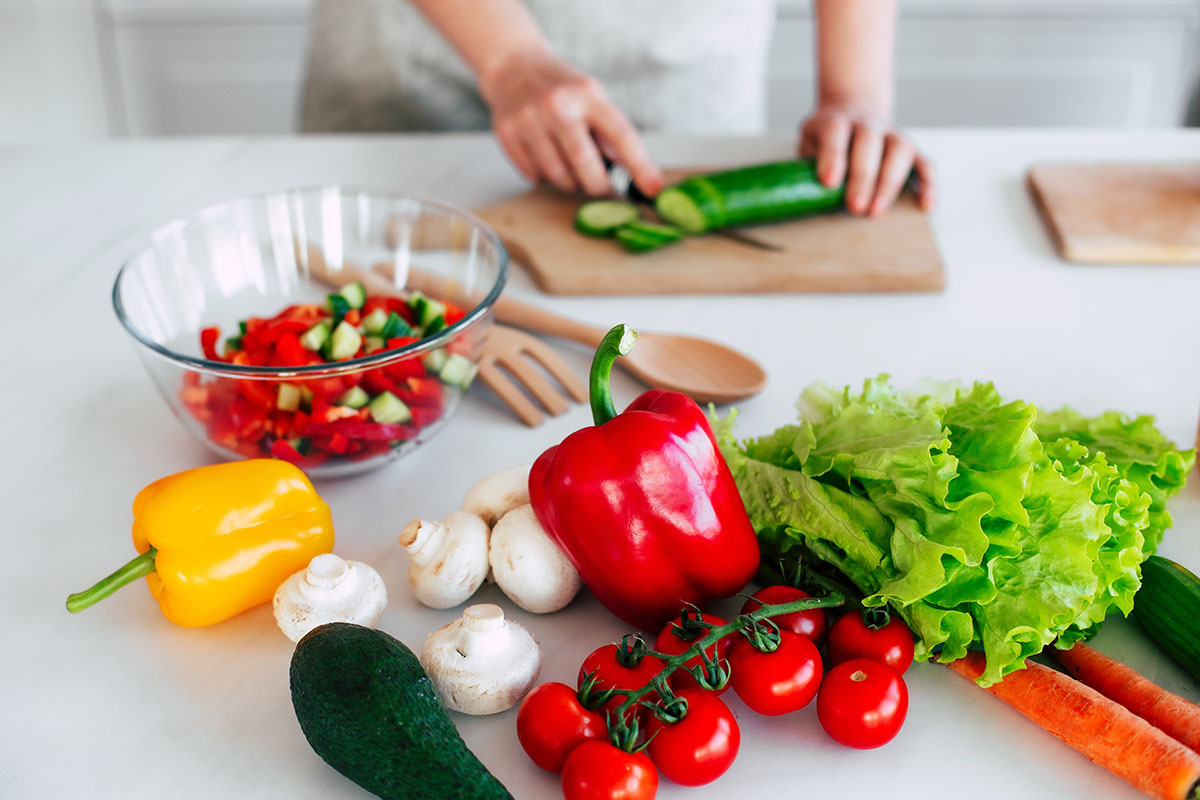 Close up photo of female hands with knife cutting fresh cucumber on the table on domestic kitchen. Many ripe, tasty and colorful vegetables with olive oil. Vegan meal, healthy nutrition