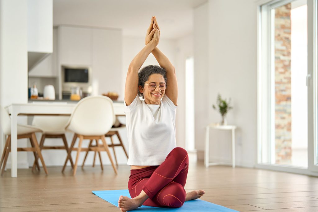 Happy healthy fit mature woman practicing yoga doing exercises sitting on mat at home interior. Sporty mindful older middle aged lady in her 50s stretching body with eyes closed in house living room.