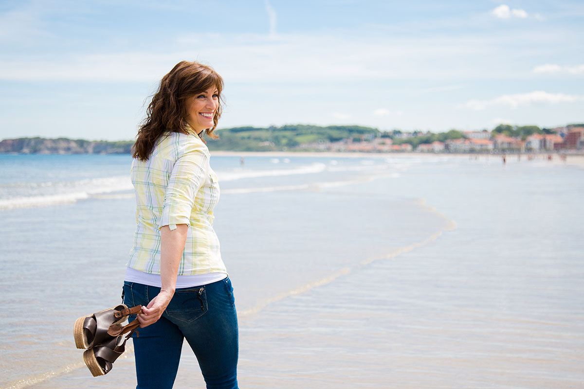 Attractive and happy middle aged woman walking along the seashore holding the shoes in hand